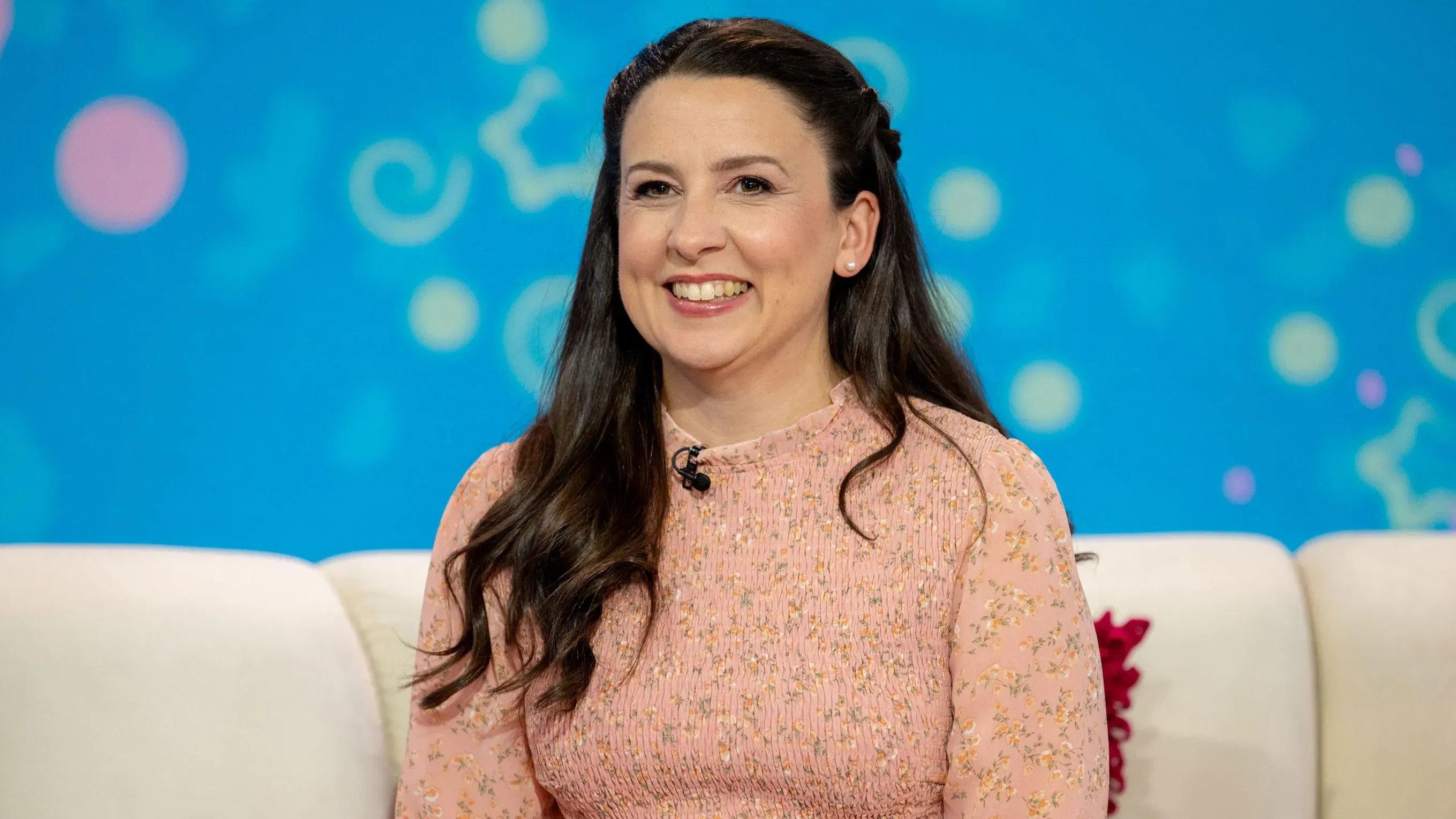 راشيل أكورسو، مدوّنة اليوتيوب المعروفة، تبتسم بينما تجلس في استوديو، مع خلفية ملونة تعكس سعادتها بمولودتها الجديدة.