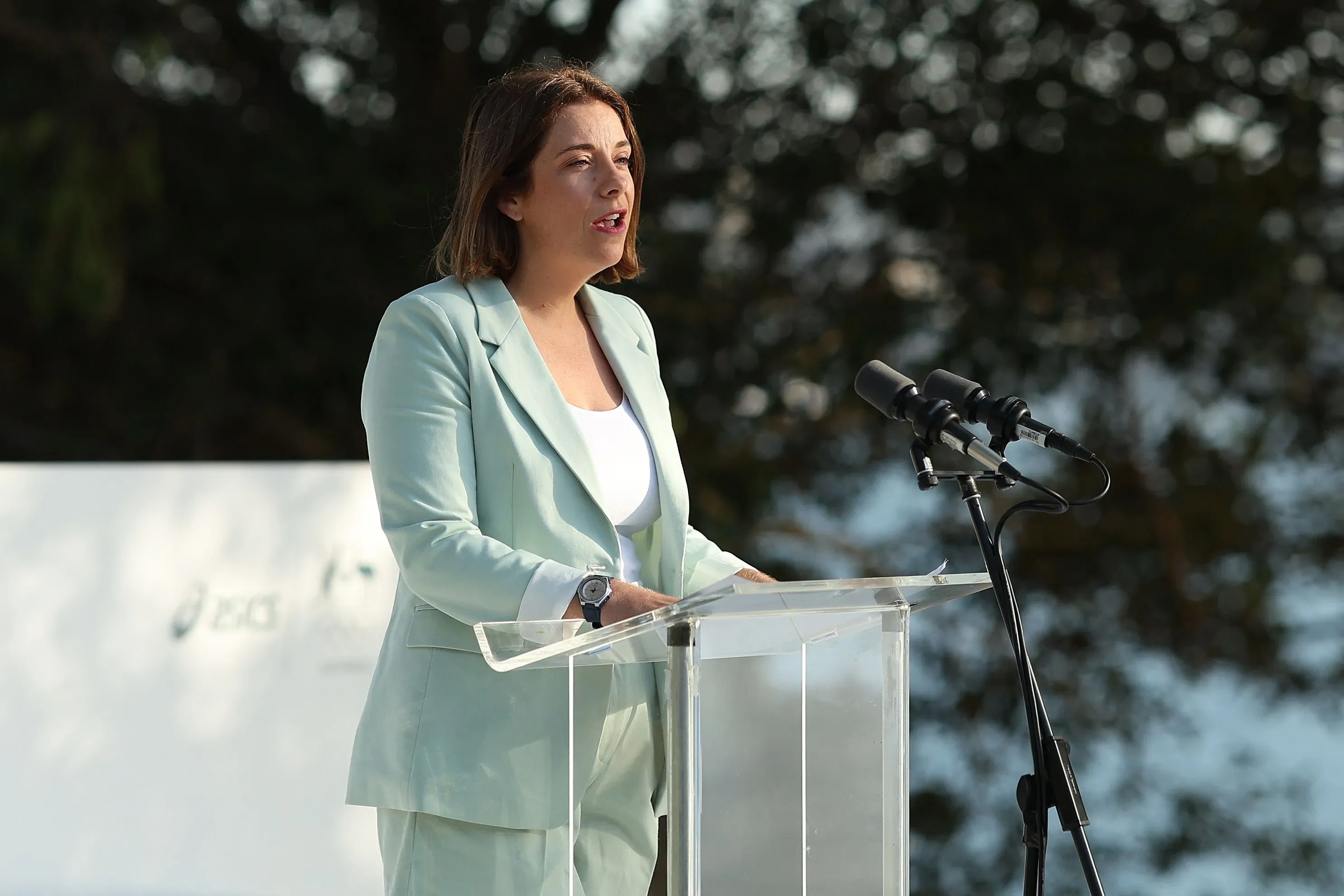 وزيرة الاتصالات الأسترالية أنيكا ويلز تتحدث أمام ميكروفونات، مع خلفية طبيعية، حول تشريع جديد لحماية الأطفال على وسائل التواصل الاجتماعي.