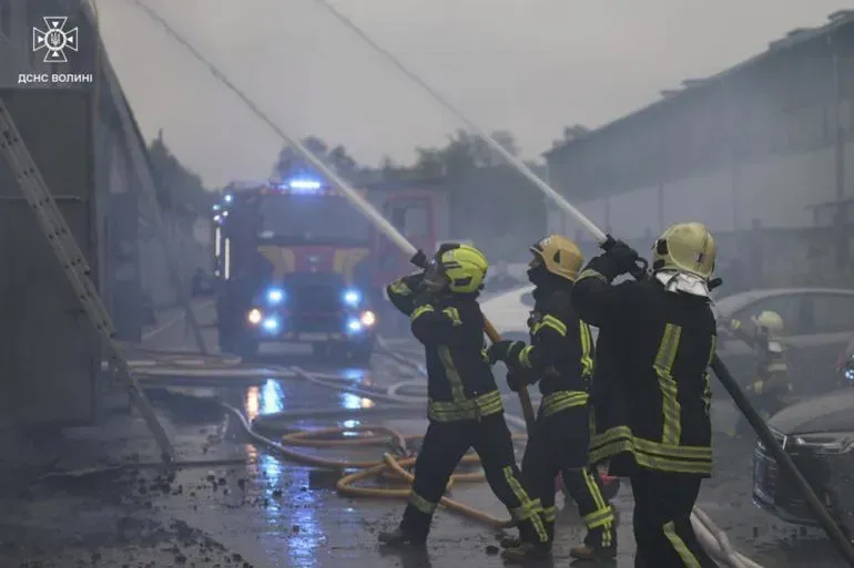 رجال إطفاء يرتدون زيًا أصفر، يستخدمون خراطيم المياه لإخماد حريق في منطقة صناعية، مع وجود شاحنة إطفاء في الخلفية وسط أجواء ضبابية.