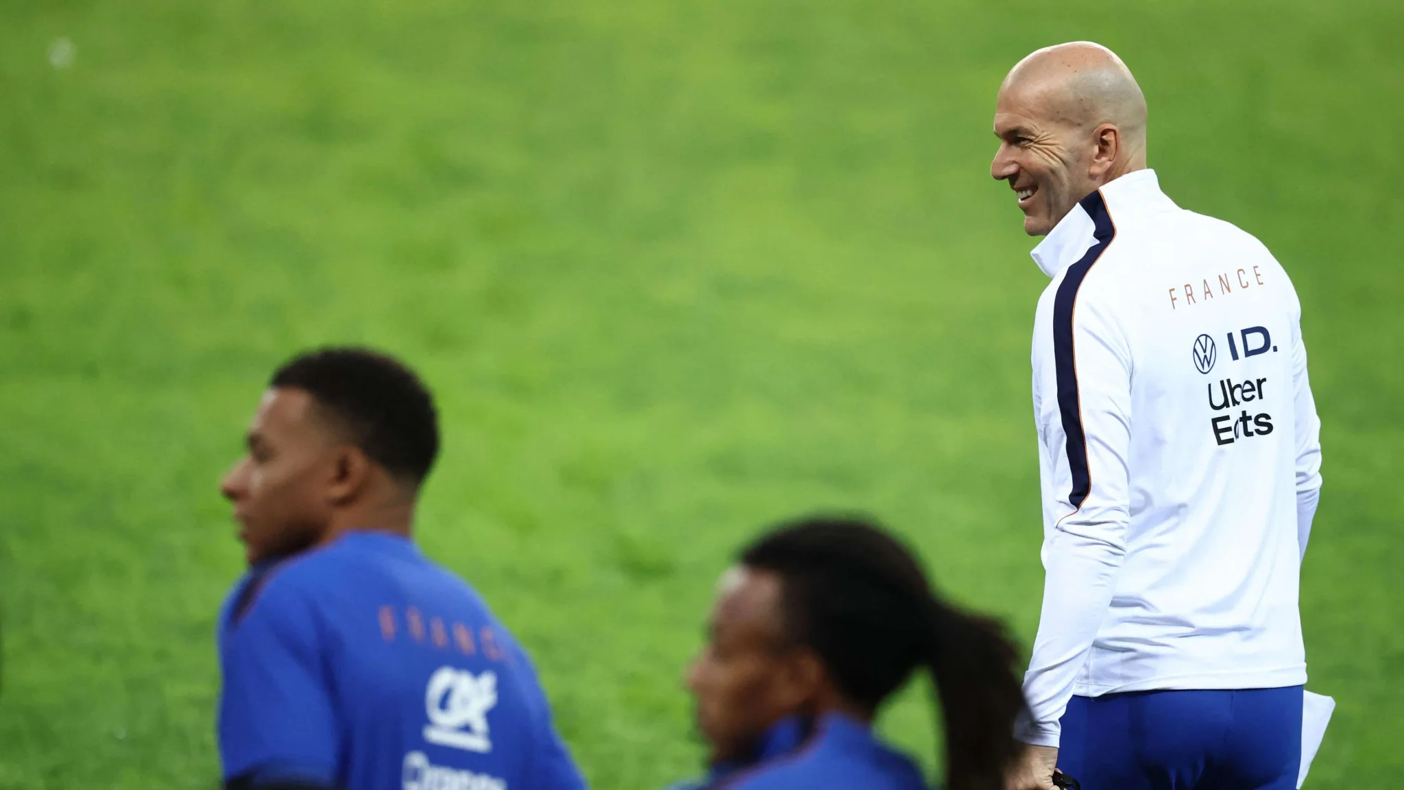 زين الدين زيدان مبتسمًا خلال تدريب منتخب فرنسا، مع وجود كيليان مبابي وزملائه في الخلفية على أرض الملعب.